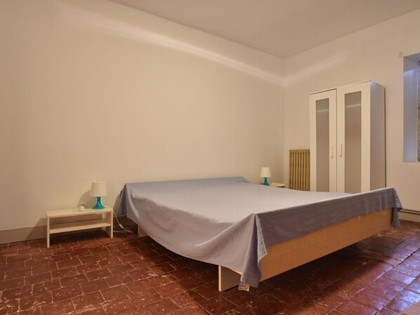 Una cama individual en una habitación con piso de ladrillo, una mesita de noche pequeña con lámpara y un armario.
