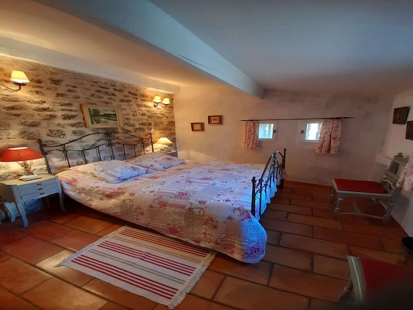 A bedroom with a stone wall, a bed with floral bedding, a nightstand with a lamp, a chair, and a window with curtains.