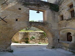 An ancient stone archway with a small window, leading to a courtyard with a stone wall and a building with a wooden door.