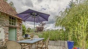 An outdoor patio with a table and chairs, a purple umbrella, and a rustic stone building in the background. - Cottage in Saint-Clair-sur-l'Elle (Saint-Clair-sur-l'Elle)