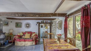 A rustic room with a wooden ceiling, a stone wall, a dining table with a patterned tablecloth, a sofa with cushions, and a window with red curtains. - Cottage in Saint-Clair-sur-l'Elle (Saint-Clair-sur-l'Elle)