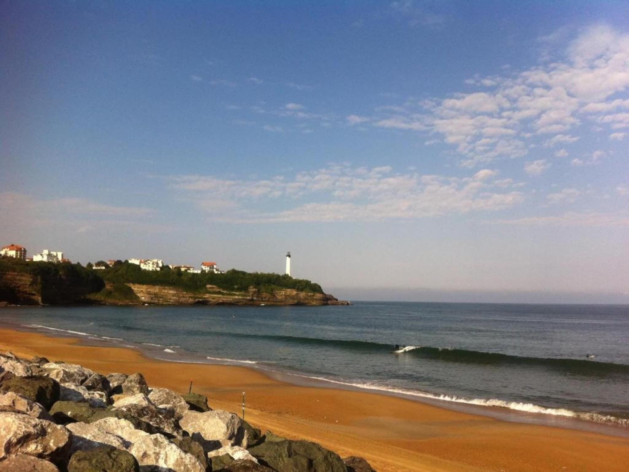 A coastal scene with a sandy beach, rocky foreground, and a lighthouse on a cliff.