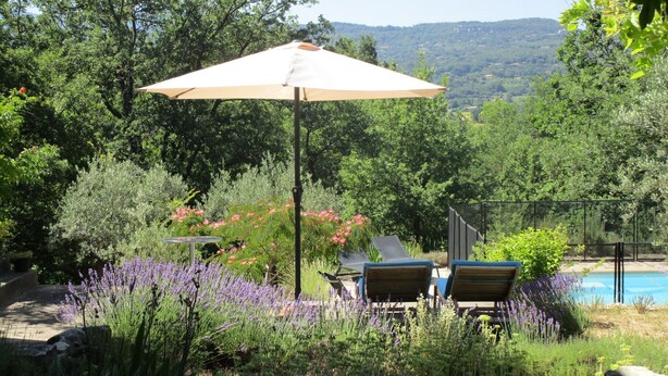 Zona de piscina con sillones de descanso y una mesa bajo una pérgola, rodeada de exuberante vegetación y vistas a las colinas.