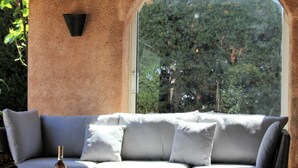 A modern outdoor seating area with a sofa and cushions, a table with a bottle, and a view of trees through a large window. - Private Pool in Provence (La Motte)