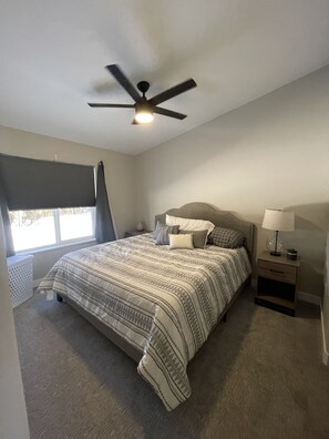 Quarto com cama, mesa de cabeceira, abajur, ventilador de teto e janela com persianas.