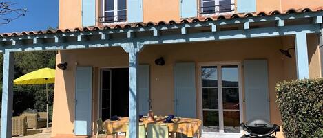 Ein zweistöckiges Haus mit überdachter Terrasse, einem für vier Personen gedeckten Esstisch und einem Grill.