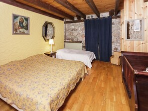 A room with a wooden ceiling, a bed with a floral bedspread, a nightstand with a lamp, a stone wall, and a window with curtains. - Charming Farmhouse in Cros-de-Géorand (Cros-De-Géorand)