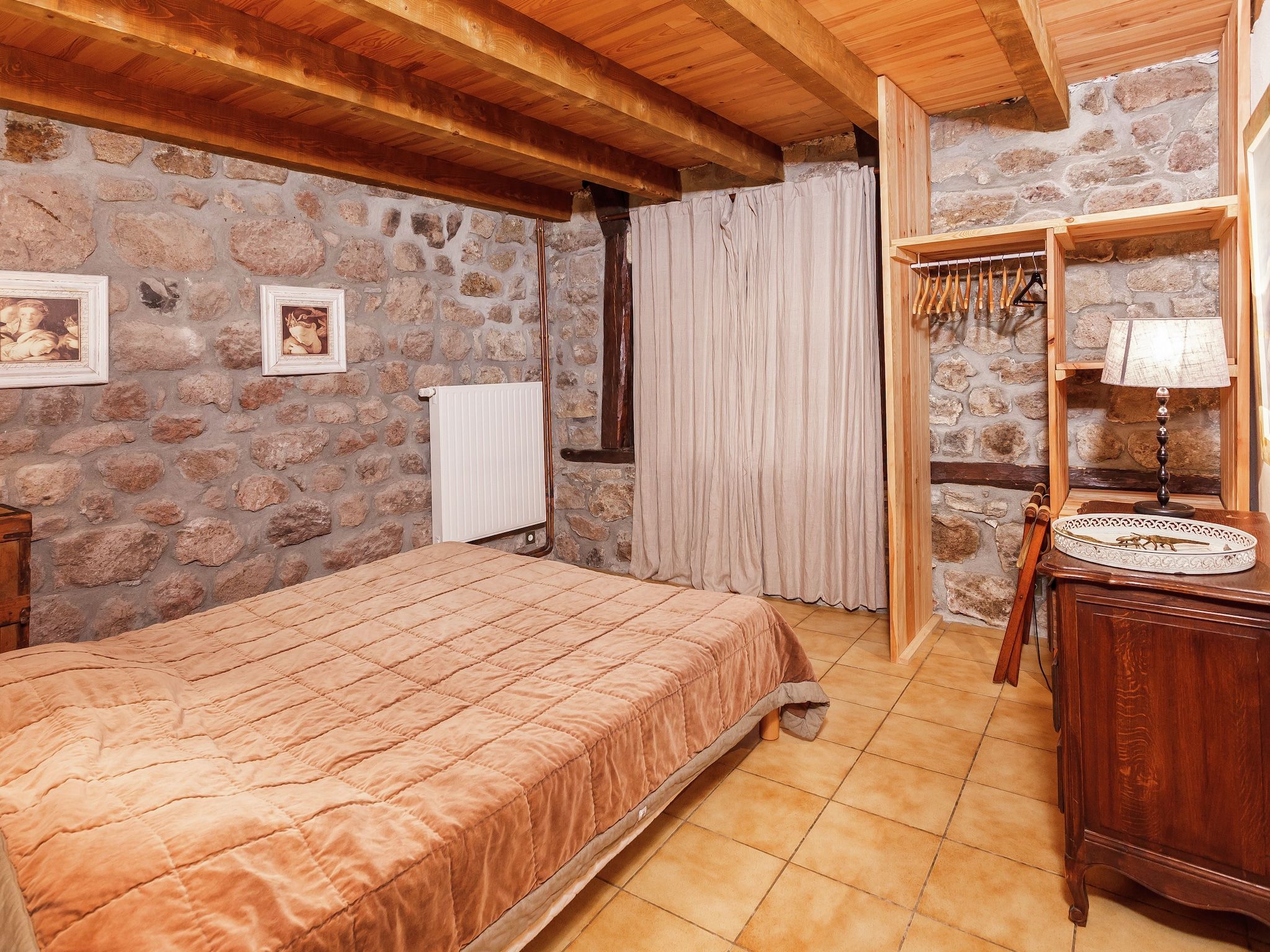 Une chambre avec un mur en pierre, des poutres en bois au plafond, un lit recouvert d’une courtepointe, un radiateur, une commode en bois et un porte-manteau suspendu.