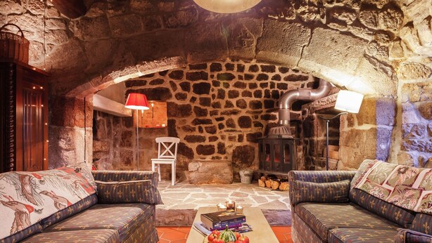 Una habitación acogedora con paredes de piedra, dos sofás estampados, una mesa de madera con libros y fruta, y una chimenea de piedra con un hogar a leña.