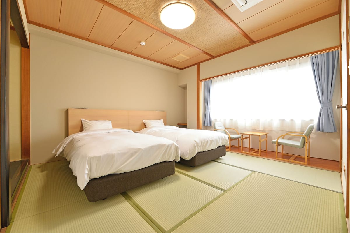 Habitación de hotel con dos camas, una mesita pequeña, una silla y un ventanal grande con cortinas.