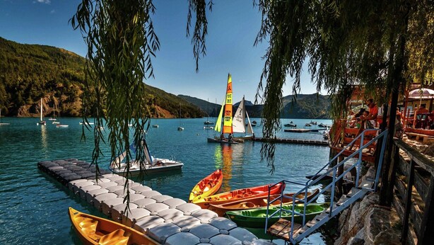 Un quai où sont amarrés des canoës et un voilier, le tout entouré d’une eau calme et de collines.