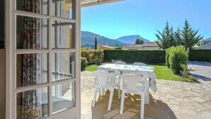 Une terrasse ouverte avec une table et des chaises, une vue sur les montagnes et un jardin agrémenté d’une haie et d’arbres.