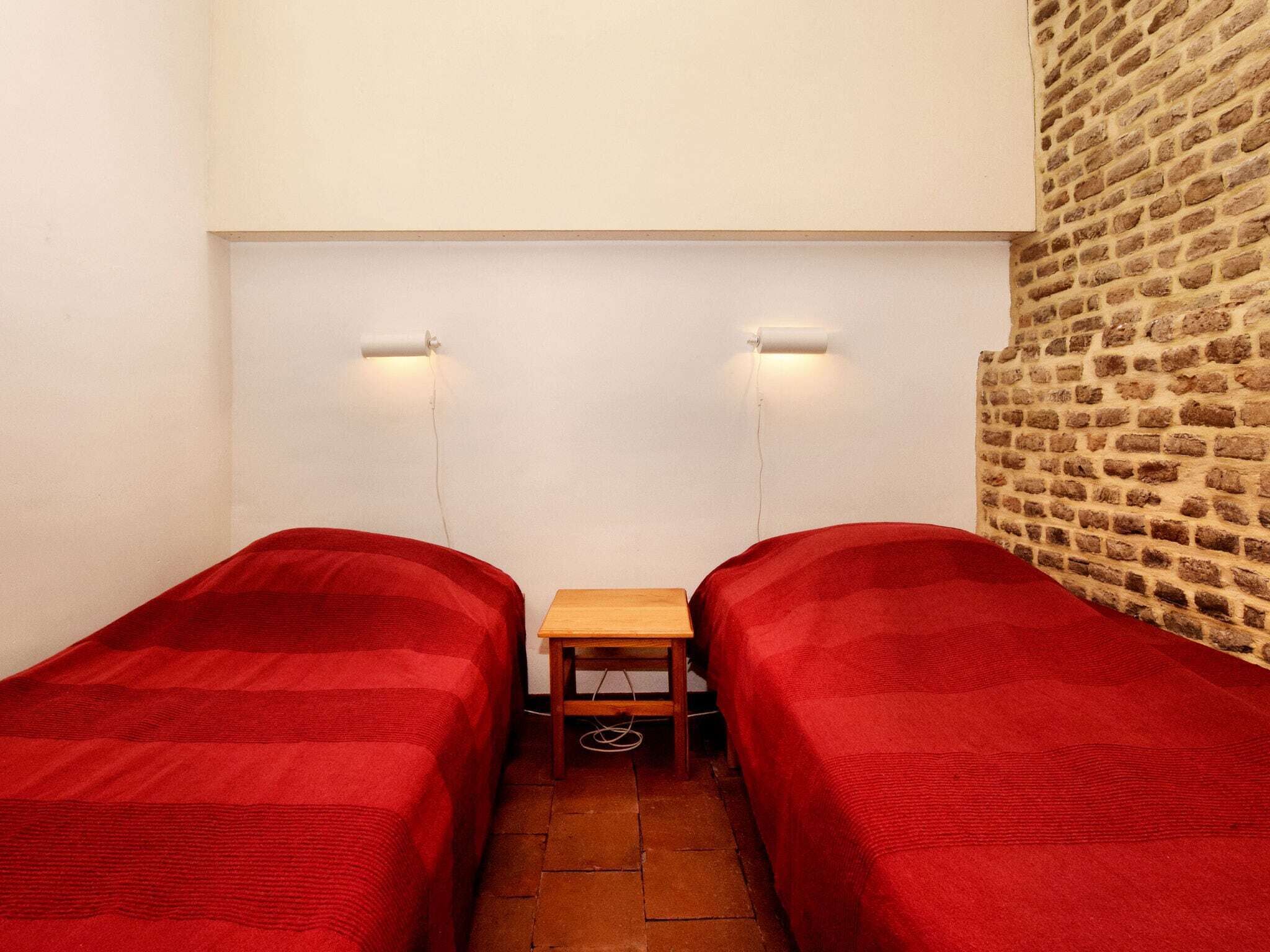 Una habitación pequeña y estrecha con dos camas individuales, cada una cubierta con un edredón rojo. En el centro hay una mesa de madera con un cable que pasa por debajo. Las paredes son lisas, y un lateral presenta un diseño de ladrillo.