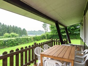 A wooden table and chairs on a covered patio with a view of a grassy area and trees. - Detached chalet with views of the lake of Butgenbach in the middle of nature (Berg)