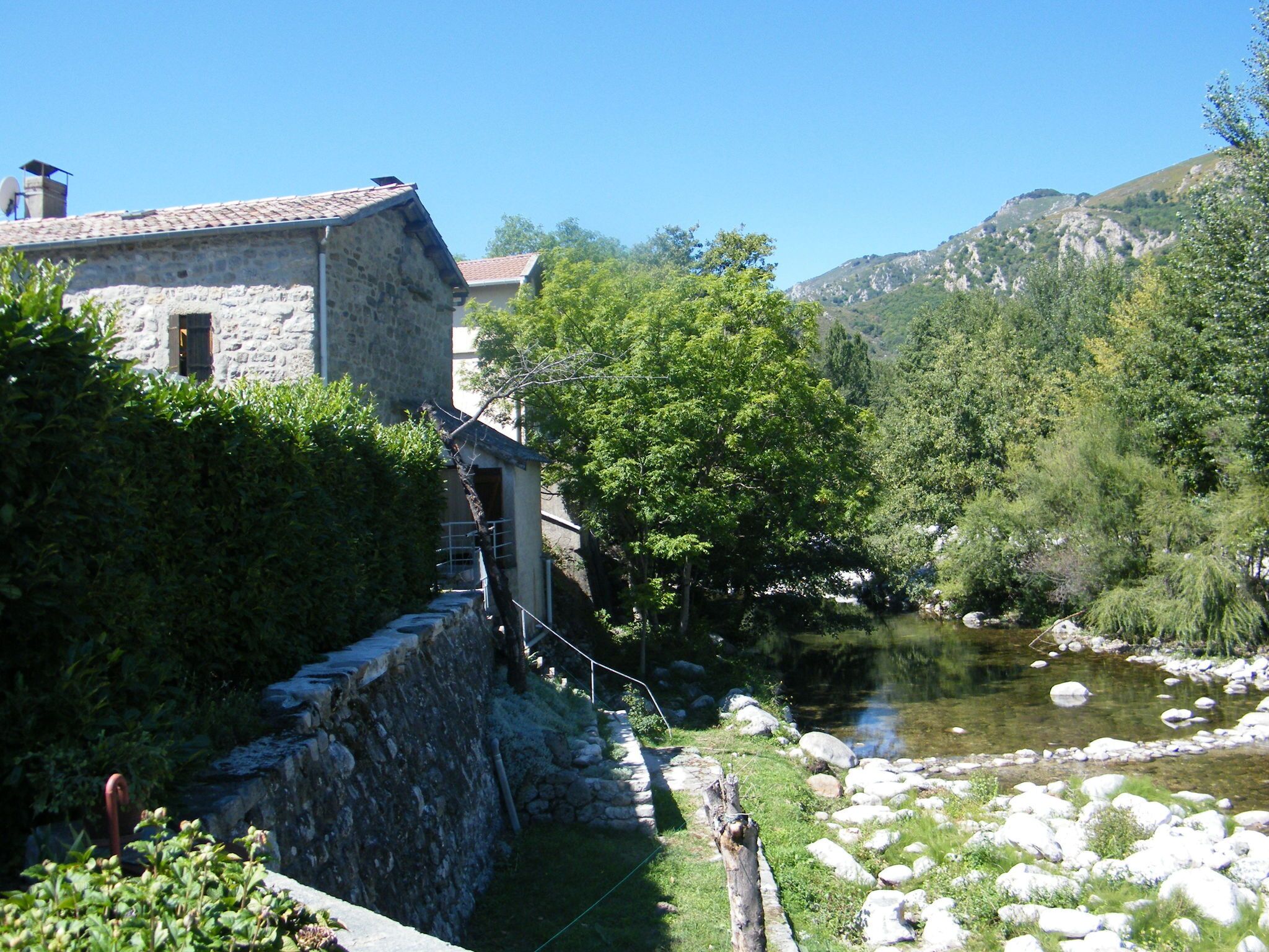 A stone building with a tiled roof, a small staircase leading to a door, and a river with rocks and greenery in the background.