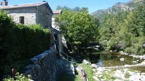 Ein Steinhaus mit Ziegeldach, eine kleine Treppe, die zu einer Tür führt, und im Hintergrund ein Fluss mit Steinen und viel Grün.