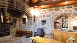 Ein Zimmer mit Steinwänden und Holzbalkendecke, ein Ledersofa, ein Holztisch mit gelber Tischdecke und ein Kamin mit einem Blumenstrauß.