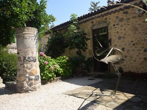 A courtyard with a stone pillar, a metal sculpture, and a building with a stone facade and a tiled roof.