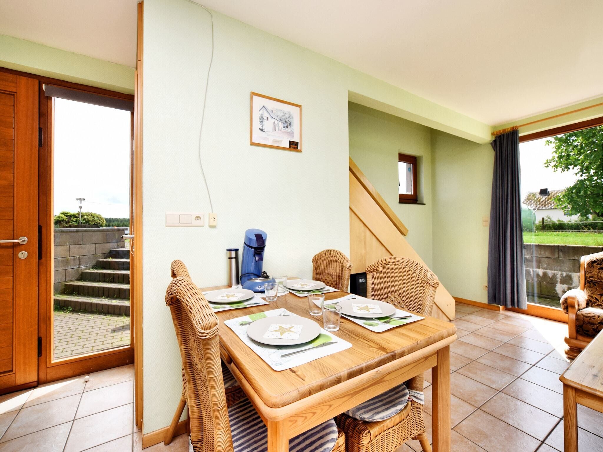 A dining area with a wooden table set for two, wicker chairs, a coffee maker, and a framed picture on the wall.