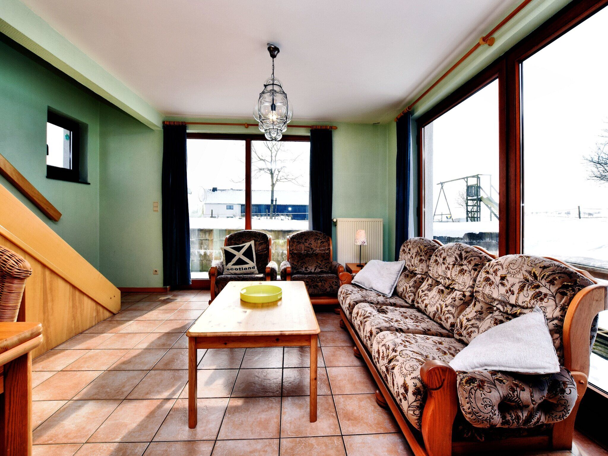 A living room with a wooden table, patterned sofa, and a view of a snowy landscape through large windows.