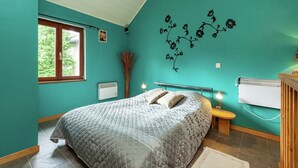 A bedroom with teal walls, a bed with a quilted grey duvet, a window with a view of greenery, a wall-mounted air conditioner, and a radiator.