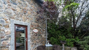 A stone building with a glass door, a sign reading "L'Orchidée", outdoor wicker furniture, and a garden with various plants.