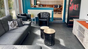 Een moderne woonkamer met een grijze bank, zwarte fauteuils en twee bijzettafeltjes met een zwart-wit patroon. In de achtergrond is een keuken te zien met een bargedeelte en een poster aan de muur.