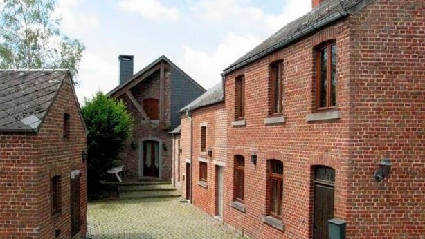 A courtyard with cobblestone paths and red brick buildings, featuring a central building with a gabled roof and a smaller building with a chimney.