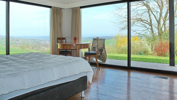 Une chambre dotée d’un grand lit, d’un bureau et d’une chaise, et offrant une vue sur un paysage verdoyant grâce à de grandes fenêtres.