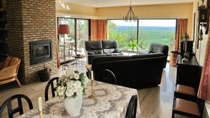 Un salon spacieux doté d’une cheminée, un coin repas avec une table dressée pour deux personnes, et une grande fenêtre offrant une vue sur l’extérieur.