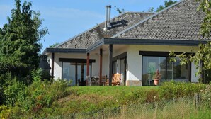 Une maison moderne avec un toit en pierre, de grandes fenêtres et un espace extérieur couvert aménagé avec du mobilier.