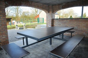 Un espace pique-nique extérieur abrité, avec une longue table et des bancs, des arches en briques, et une vue sur une aire de jeux et des bâtiments en arrière-plan.