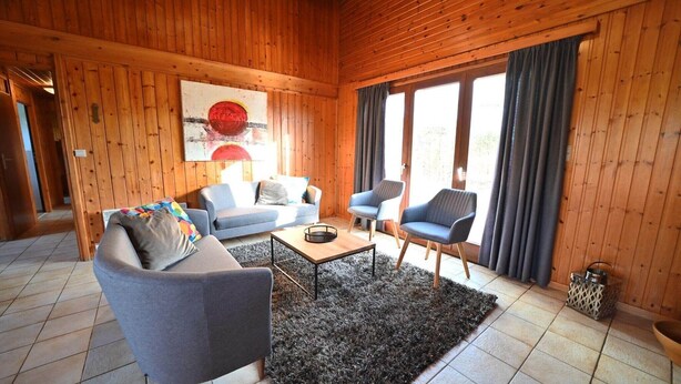 Ein gemütliches Holzhütten-Interieur mit einer Couch, Sesseln, einem Couchtisch und einem großen Fenster mit Vorhängen.