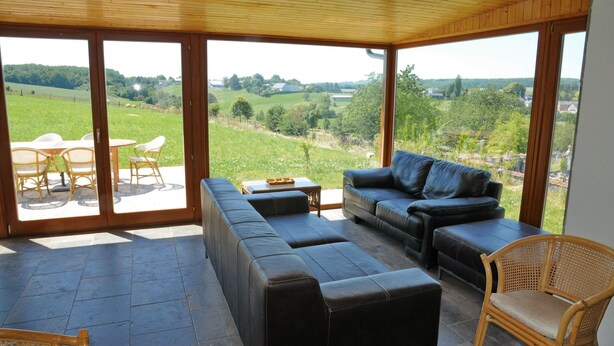 A sunlit patio with a sofa, chairs, and a table, overlooking a green landscape. - Stone House in Durbuy with Sauna & Garden Views (Bonsin)