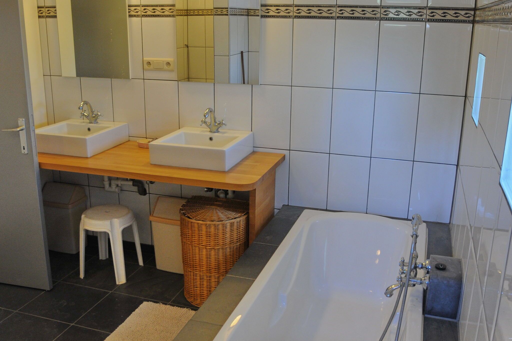 Een badkamer met twee wasbakken, een bad, een spiegel, een krukje en een rieten mand.