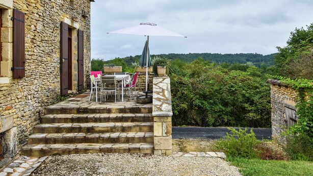 Un bâtiment en pierre avec un escalier extérieur menant à une table et des chaises placées sous un grand parasol.