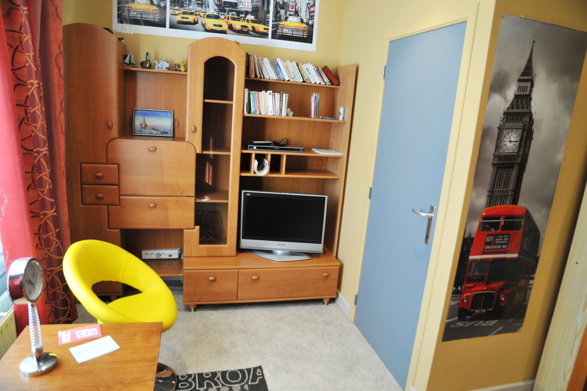 A room with a wooden entertainment unit, a television, and a door with a poster of a red double-decker bus.