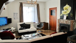 Un salon avec un canapé blanc, un fauteuil noir et une table basse. Il y a un téléviseur à gauche, une cheminée à droite et une fenêtre avec des rideaux.