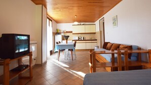 Una stanza accogliente con soffitto in legno, una televisione su un mobile, un tavolo da pranzo con sedie, un divano e un angolo cottura sullo sfondo.
