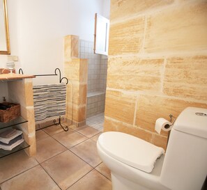 Un baño con pared de piedra, inodoro blanco, un perchero con toalla a rayas, un estante pequeño con una cesta y un espejo.