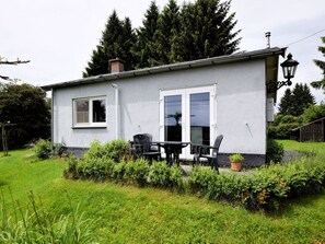 Ein kleines, eingeschossiges Haus mit weißer Fassade, einer Haustür und einem Fenster. Vor der Tür befindet sich eine Terrasse mit Tisch und Stühlen sowie ein Blumentopf.