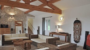 A spacious living area with wicker furniture, a stone wall, wooden beams, and a staircase leading to another level. - Cosy holiday home with views and private pool (Saint-Ambroix)