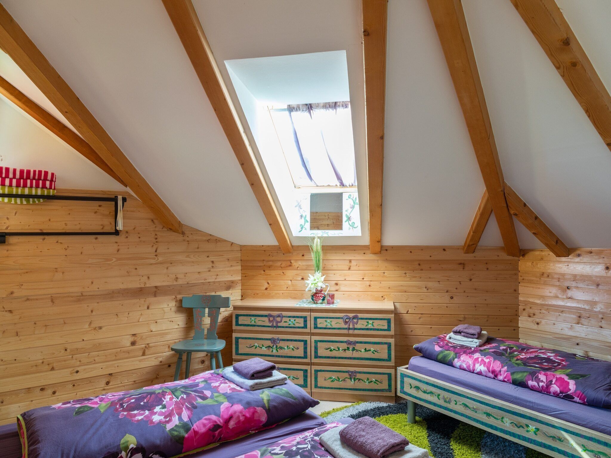 A cozy attic bedroom with wooden walls, a skylight, two beds with floral bedding, a dresser, and a chair.