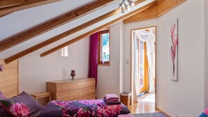 Een slaapkamer met een schuine kap, houten balken, een bed met bloemenbeddels, een dressoir en een schilderij van bloemen.