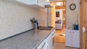 Een moderne keuken met granieten aanrechtbladen, witte kasten en roestvrijstalen apparatuur. Achtergrond toont een eethoek met een houten plafond en een knusse sfeer.