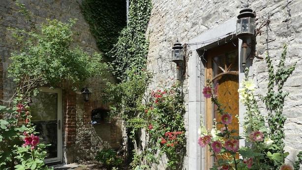 Un edificio de piedra con puerta de madera, un sendero de ladrillo y diversas plantas y flores.