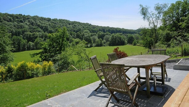 Une table et des chaises en bois sur une terrasse dotée d’un sol en pierre, offrant une vue sur une vallée verdoyante et des collines boisées.