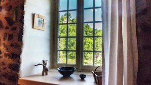 Una habitación con un ventanal grande, una pared de piedra, un estante pequeño con un cuenco y una figurita, y un cuadro en la pared.
