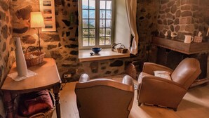 Una habitación acogedora con paredes de piedra, una chimenea, una ventana con cortinas, una mesa con una lámpara y dos sillones de cuero.