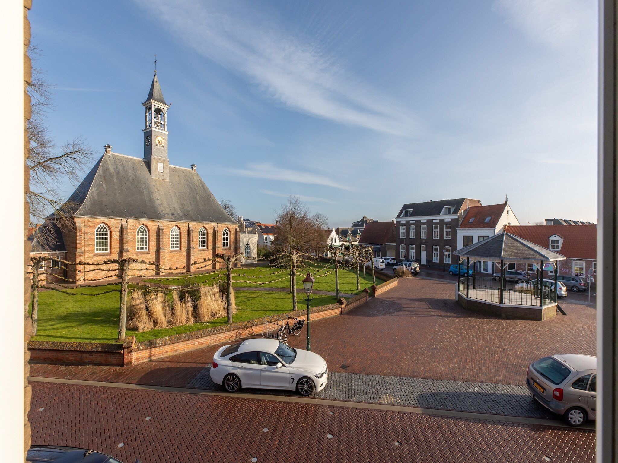 Une église dotée d’un clocher, une place pavée avec des voitures, et des bâtiments traditionnels en arrière-plan.